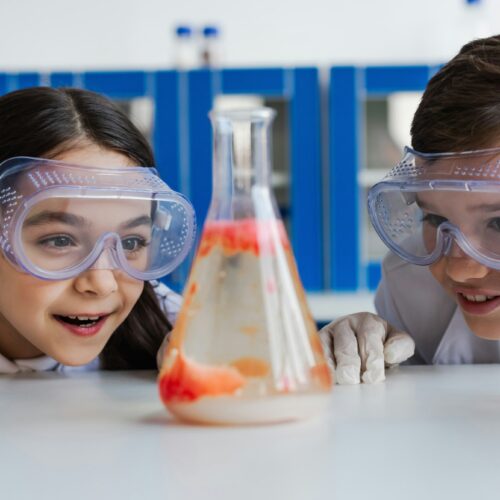 amazed kids in goggles looking at blurred flask with liquid while doing chemical experiment in lab